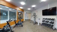 This fitness center is a purpose-built exercise facility designed to support a variety of workout routines. The space is bright and clean, featuring white walls accented with bold orange framing around interior windows that overlook an adjacent area, adding an open and connected feel to the room. The flooring is a durable speckled carpet-tile surface well-suited for gym use. Equipment includes a stationary cycling bike for cardiovascular training, a multi-function cable resistance machine ideal for a range of strength exercises, and a tiered dumbbell rack stocked with free weights for targeted muscle training. A wall-mounted flat-screen television is positioned for easy viewing during workouts. Recessed and flush-mounted ceiling lights provide consistent, even illumination throughout the space. Glass doors at the rear of the room offer transparency and connectivity to the surrounding facility. A security camera and fire alarm are also present, reflecting a commitment to safety. Overall, the fitness center offers a clean, functional, and well-organized environment suitable for both cardio and strength training needs.