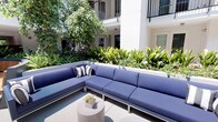 This outdoor courtyard is nestled within the interior of a modern, multi-storey residential complex, surrounded on multiple sides by white-rendered building facades adorned with black-framed balconies. The design creates a sheltered, semi-private atmosphere while remaining fully open to natural light and fresh air.

The courtyard is organized into distinct zones that encourage both relaxation and social interaction. A generously sized L-shaped sectional sofa upholstered in deep blue is positioned alongside raised garden beds brimming with lush tropical foliage, including broad-leafed plants and layered ground cover. This seating area is backed by a canopy of mature trees, providing natural shade and a sense of enclosure.

Further into the space, a curved timber bench wraps around a large cylindrical planter containing tall ornamental grasses and palms, creating a natural focal point. Sleek charcoal-toned tables are paired with woven rattan egg chairs, offering a more intimate setting for smaller gatherings.

The flooring throughout is a smooth, light-toned concrete surface that complements the clean, contemporary aesthetic of the surrounding architecture. Raised planter walls with integrated low-level lighting add definition to the pathways while enhancing the ambiance during evening hours. Overall, the courtyard offers a well-appointed, resort-inspired outdoor environment.