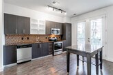 This single-floor space offers a well-designed open-concept layout that seamlessly connects the kitchen and living area. The kitchen is outfitted with dark espresso cabinetry, including frosted glass upper cabinet inserts, and is complemented by rich dark granite countertops and a mosaic tile backsplash in warm brown tones. Stainless steel appliances, including a range, microwave, and dishwasher, add a contemporary edge, while a compact island provides additional workspace and a casual dining option. Track lighting illuminates the kitchen efficiently.

Dark hardwood floors extend throughout the main living area, creating a cohesive and polished aesthetic. A hallway leads to the bedroom and bathroom, where carpet flooring adds warmth to the sleeping space. The bathroom features light tile flooring and a glass-enclosed walk-in shower, with direct access to a closet equipped with a clothing rod. French doors and adjacent windows in the main living area invite natural light and provide views of the surrounding greenery and cityscape, enhancing the overall brightness and openness of the space.