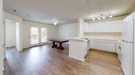 Located at 801 S German Ln in Conway, Arkansas, this one-bedroom, one-bathroom unit is thoughtfully laid out across a single floor. The heart of the home is a combined living, dining, and kitchen space that feels open and connected. The kitchen features white cabinetry, a peninsula with a built-in sink, track lighting, and white appliances including a range and refrigerator — all set against light hardwood-style flooring that runs throughout the unit. The dining and living area is bathed in natural light, courtesy of a set of French doors that open to the outside, complemented by a ceiling fan overhead. The bedroom is a comfortable, well-lit room with a large window offering views of surrounding greenery and includes access to a walk-in closet, providing ample storage. Additional closets further enhance the unit's storage capacity. The bathroom is designed with accessibility in mind, featuring an open vanity with a mirror, grab bars, and hardwood-style flooring consistent with the rest of the unit. A dedicated utility room rounds out the layout. The overall aesthetic is clean and neutral, with light walls and warm flooring creating a bright, inviting atmosphere throughout.
