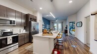 This thoughtfully designed 1-bedroom, 1-bathroom residence features an open-concept floor plan that seamlessly connects the kitchen, dining, and living areas. The kitchen is a standout, boasting dark espresso cabinetry, granite countertops, a large center island with bar seating, stainless steel appliances including a French door refrigerator and built-in microwave, and a dedicated wine rack. Rich hardwood-style flooring runs throughout the main living areas, anchoring the space with warmth and continuity. The living room is highlighted by a bold blue accent wall, a ceiling fan, and access to natural light through large windows and a glass door. The bedroom is generously sized, featuring high windows for natural light, a ceiling fan, plush carpeting, and its own striking blue accent wall. A dedicated walk-in closet offers ample storage with multiple hanging rods and built-in shelving. The bathroom features a granite-topped double vanity with dark cabinetry, large-format tile flooring, and a clean, contemporary aesthetic. Recessed lighting and modern pendant fixtures throughout the space enhance its polished, urban feel.