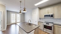 This single-floor one-bedroom unit offers a thoughtfully designed layout that maximizes functionality and style. The open-concept kitchen and dining area serves as the heart of the space, featuring a central island with a dark stone countertop, an undermount sink, and a brushed gold faucet. Pendant lights with brass finishes hang above the island, adding a warm, modern touch. The kitchen is equipped with stainless steel appliances — including a range, built-in microwave, and refrigerator — complemented by light wood cabinetry and a classic white subway tile backsplash. Wood-look flooring runs throughout the kitchen and dining area, which also benefits from natural light through large windows and a glass door that opens to a balcony or exterior space. The bedroom offers a cozy atmosphere with soft carpeting, a ceiling fan, and a window with outdoor views. Neutral gray tones on the walls throughout the unit create a clean, cohesive aesthetic. A dedicated walk-in closet with wire shelving provides practical storage. The overall design balances contemporary finishes with a comfortable, livable feel.