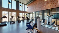 Located at 2439 Beatrice St in Dallas, TX, this single-floor fitness center is designed to deliver a professional-grade workout environment with a modern, industrial aesthetic. The space is distinguished by its dramatically tall ceilings, which lend an open, airy atmosphere throughout the facility. Large, floor-to-ceiling windows line the walls, flooding the interior with abundant natural light and offering views of the surrounding outdoor environment.

The main workout area is generously proportioned, providing ample space between equipment stations for comfortable use. A standout architectural feature is the textured, patterned accent wall finished in warm terracotta tones, adding visual interest and a distinctive design identity to the space. Pendant lighting with dark industrial-style fixtures complements the overall aesthetic.

The facility is outfitted with a comprehensive selection of commercial-grade equipment, including treadmills, ellipticals, stair climbers, stationary bikes, and a wide array of Hoist and True strength machines. A dedicated section houses cable and resistance machines, while additional storage cubbies keep the space organized. Dark rubber flooring throughout provides durability and safety. Glass partition walls separate distinct workout zones while maintaining an open, connected feel across the entire facility.