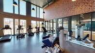 Located at 2439 Beatrice St in Dallas, TX, this single-floor fitness center is designed to deliver a professional-grade workout environment with a modern, industrial aesthetic. The space is distinguished by its dramatically tall ceilings, which lend an open, airy atmosphere throughout the facility. Large, floor-to-ceiling windows line the walls, flooding the interior with abundant natural light and offering views of the surrounding outdoor environment.

The main workout area is generously proportioned, providing ample space between equipment stations for comfortable use. A standout architectural feature is the textured, patterned accent wall finished in warm terracotta tones, adding visual interest and a distinctive design identity to the space. Pendant lighting with dark industrial-style fixtures complements the overall aesthetic.

The facility is outfitted with a comprehensive selection of commercial-grade equipment, including treadmills, ellipticals, stair climbers, stationary bikes, and a wide array of Hoist and True strength machines. A dedicated section houses cable and resistance machines, while additional storage cubbies keep the space organized. Dark rubber flooring throughout provides durability and safety. Glass partition walls separate distinct workout zones while maintaining an open, connected feel across the entire facility.