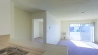 This single-floor residence offers a straightforward and functional layout designed for comfortable everyday living. The interior is characterized by neutral white walls and a textured popcorn ceiling that runs throughout, lending a consistent and cohesive aesthetic to the space.

The kitchen area features tiled countertops and flooring, providing a practical and easy-to-maintain surface for meal preparation. A sink is conveniently positioned within view, and the open sightlines from the kitchen extend directly into the adjacent living areas.

The living space is finished with plush carpeting in a soft purple-grey tone, creating a warm and inviting atmosphere underfoot. A wall-mounted air conditioning unit is positioned near the exterior wall, ensuring climate control comfort year-round. The room benefits greatly from a large sliding glass door that opens to an outdoor area, flooding the interior with abundant natural light and providing a seamless connection to the outside.

A doorway leads to an additional room, also finished with carpeting and white walls, suitable for use as a bedroom or flex space. The overall design of the residence is clean and neutral, offering a blank canvas that is well-suited for personalization. The single-floor plan ensures easy accessibility and convenient flow between all areas of the home.