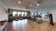 This single-floor fitness center is thoughtfully designed to support a wide range of workout routines. The main exercise area features hardwood-style flooring throughout, recessed lighting, and a ceiling fan that contribute to a bright and comfortable training environment. A row of windows along one wall allows natural light to fill the space, creating an open and energizing atmosphere.

The cardio zone is well-stocked with multiple treadmills and elliptical machines, complemented by wall-mounted televisions for entertainment during workouts. A multi-station cable machine and power tower provide options for comprehensive strength training, while large wall mirrors reflect the space and assist with form correction. A rowing machine adds further versatility to the cardio offerings.

A dedicated functional training area features rubber mat flooring, a freestanding Century punching bag, medicine balls, stability balls, a weight bench, and yoga mats, making it ideal for stretching, core work, and dynamic exercises. Motivational fitness posters adorn the walls throughout this zone.

Additional amenities include dual wall-mounted water fountains, ensuring hydration is always convenient. The space is clean, well-maintained, and efficiently organized, offering everything needed for a complete fitness experience in a polished and functional setting.