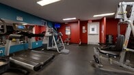 Located at 2911 Bayview in Toronto's M2K neighbourhood, this fitness center offers a well-equipped workout space across a single floor. The room makes a strong visual impression with its bold red and teal accent walls, dark rubber flooring, and large wall mirrors that enhance the sense of space and allow users to monitor their form during exercise.

The cardio section is anchored by Life Fitness treadmills and elliptical trainers, providing a solid range of aerobic options. A dedicated strength training area features a multi-station cable machine, a weight bench, and a rack of dumbbells in varying sizes, supporting a full range of resistance exercises. A collection of medicine balls and stability balls rounds out the functional fitness offerings, complemented by wall-mounted exercise guide charts for reference.

Overhead, flush-mounted fluorescent panel lighting ensures the space is evenly and brightly lit throughout. A wall-mounted flat-screen television adds an entertainment option for cardio sessions. An adjacent anteroom with seating provides a convenient area for rest or transitions. The facility is equipped with a water station and is accessible via a keyed entry door, ensuring a secure and comfortable environment for users.
