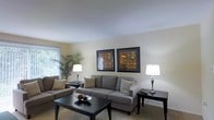 This single-floor residence offers a thoughtfully designed living space with a clean, neutral color palette that creates a calm and welcoming atmosphere. The living room serves as the centerpiece of the home, featuring plush gray upholstered sofas arranged around a dark-finished rectangular coffee table, ideal for both relaxing and entertaining. Soft carpeted flooring adds warmth underfoot, while the smooth, light-colored walls provide a bright and airy backdrop throughout the space. Natural light filters in through a window dressed with vertical blinds, complementing the warm glow cast by classic table lamps placed on matching side tables. A pair of bold, framed wall art pieces with rich earth-tone geometric patterns adds a decorative focal point to the main wall, contributing visual interest without overwhelming the room's understated elegance. A lush potted palm plant in the corner introduces a touch of nature, enhancing the fresh and livable feel of the interior. The overall design aesthetic is modern and practical, with carefully chosen furnishings and accessories that balance style and comfort, making this a functional and inviting single-level living space.