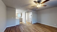 This single-floor space combines practical design with modern updates throughout. The living area is open and well-proportioned, featuring warm wood-look plank flooring, neutral gray walls, and a textured ceiling with a ceiling fan that provides both light and airflow. Natural light enters through blinded windows, offering views of surrounding greenery. The space flows seamlessly toward the kitchen and additional rooms, creating a connected, functional layout.

The kitchen is thoughtfully designed with medium-toned wood cabinetry on both upper and lower levels, complemented by a vertical tile backsplash and white quartz countertops with subtle veining. A peninsula provides additional prep and seating space. Stainless steel appliances, including a gas range, over-range microwave, and refrigerator, complete the setup alongside a single-basin sink with a modern faucet beneath a window.

The bathroom is clean and well-appointed, featuring a wood-toned vanity with a white countertop, a backlit mirror, chrome fixtures, and a full tub-shower combination with a bright white surround. Flooring continues the wood-look planks found throughout the unit. The overall ambiance is bright, neutral, and move-in ready, reflecting a cohesive and updated aesthetic.