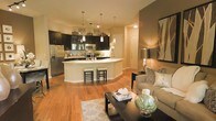 This single-floor space showcases an open-concept layout that seamlessly integrates the kitchen and living area into one cohesive, functional environment. The kitchen is a standout feature, boasting dark espresso cabinetry that contrasts sharply against the bright white curved island, which doubles as a breakfast bar with seating for multiple guests. Stainless steel appliances, including a French door refrigerator and built-in microwave, complement the space, while a mosaic tile backsplash adds a refined decorative touch. Pendant lighting with crystal-style fixtures hangs above the island, providing both task lighting and visual interest.

Warm hardwood flooring runs throughout, unifying the kitchen and living areas and contributing to the overall inviting ambiance. The living area is thoughtfully arranged with neutral-toned furnishings, a patterned area rug, and a sleek dark-framed coffee table. An accent wall in a deeper earth tone anchors the living space and provides a rich contrast to the lighter surroundings. Track lighting and recessed ceiling fixtures ensure the entire space is well-lit, enhancing its open and airy feel throughout.