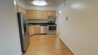 This single-floor space centers around a well-appointed kitchen that serves as a focal point of the layout. The kitchen is equipped with a full suite of stainless steel appliances, including a French door refrigerator, electric range, and dishwasher, offering a modern and cohesive look. Light maple cabinetry lines the walls, providing generous storage space above and below the dark countertops, which create a pleasing contrast against the lighter tones of the room. An under-cabinet range hood sits above the stove, adding a practical element to the cooking area.

The flooring throughout is light hardwood-style, lending warmth and continuity to the space. Crown molding along the ceiling adds a subtle architectural detail, while a flush-mount ceiling light fixture provides consistent overhead illumination. The walls are painted in a clean, neutral white, keeping the space feeling open and bright. A doorway visible from the kitchen suggests additional rooms beyond, contributing to a functional and connected floor plan. The overall ambiance is clean, practical, and move-in ready.