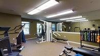 This single-floor fitness space is designed to support a well-rounded workout routine in a compact yet functional environment. The room is outfitted with a multi-station cable weight machine, a workout bench, and additional strength and cardio equipment, providing users with a variety of exercise options. The flooring is a neutral carpet, offering a comfortable surface throughout the space.

A large wall mirror spans one side of the room, visually expanding the area and allowing users to monitor their form during exercise. The walls are painted in an earthy olive green tone, lending the space a grounded, utilitarian feel. Overhead, recessed fluorescent light fixtures provide consistent, even illumination throughout the room.

Natural light filters in through a window on one wall, offering a view of the surrounding greenery and adding a touch of brightness to the interior. The textured ceiling completes the space's straightforward, no-frills aesthetic. Overall, the fitness room is a practical, well-equipped space suited for daily exercise needs.