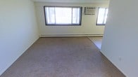 This single-floor space features an open living area finished with wall-to-wall carpeting that transitions into a tiled section, offering a practical and functional layout. The room is illuminated by natural light streaming through a large multi-pane window fitted with vertical blinds, complemented by an additional side window that further brightens the interior. Climate control is provided by a wall-mounted air conditioning unit alongside a baseboard heating system that runs along the perimeter of the room, ensuring year-round comfort. The neutral, light-colored walls contribute to a clean and open atmosphere, making the space feel airy despite its modest proportions. The transition from carpet to tile near the corner suggests a defined boundary between living and utility or entryway areas, adding a degree of functional zoning to the layout. Overall, the space presents a straightforward, no-frills interior that offers a practical foundation for a variety of uses.