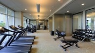 This fitness center offers a comprehensive workout environment designed to accommodate a wide range of exercise needs. The space is laid out on a single floor, with a generous open layout that allows for easy movement between equipment zones. Along one side, a row of cardio machines — including treadmills and ellipticals — lines the wall beneath large windows that invite natural light and provide views of the surrounding greenery. The neutral-toned walls and patterned carpet flooring create a clean, modern aesthetic throughout. Recessed ceiling lighting, complemented by pendant-style fixtures, ensures the space is consistently and evenly illuminated. On the opposite side, strength and flexibility training equipment is arranged to maximize usability, including an adjustable weight bench and additional free-standing machines. A mirrored wall at the far end of the room visually expands the space and supports form-checking during workouts. Exercise balls and supplementary fitness accessories are neatly stored within reach. The overall ambiance is calm, professional, and well-organized, making this fitness center a functional and inviting space suited for both casual users and dedicated fitness enthusiasts.
