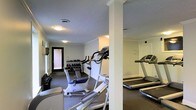 This fitness center offers a practical and well-organized space designed to support a range of workout routines. The room is finished with crown molding and a textured ceiling, lending a polished, classic character to the utilitarian space. Structural columns divide the floor plan into distinct workout zones without compromising the overall openness of the area.

The cardio section is well-stocked, featuring multiple treadmills and an elliptical trainer positioned to allow comfortable use. A dedicated free weights area lines one wall, with a tiered dumbbell rack offering a full range of weights for strength training exercises. The dark, low-pile carpet throughout provides a stable and comfortable surface underfoot.

Overhead flush-mount lighting ensures the entire space is evenly and adequately illuminated. A glass-panel entry door at one end of the room allows natural light to filter in from an exterior brick-accented entryway, adding a welcoming touch. The layout is efficient and easy to navigate, making the most of the available floor space. Overall, the fitness center presents a clean, functional, and inviting environment well-suited for daily exercise.