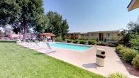 This outdoor community space is centered around a generously sized rectangular swimming pool with clear, blue water, framed by a wide concrete pool deck lined with white lounge chairs for relaxed sunbathing. A handsome wooden pergola provides a shaded gathering area adjacent to the pool, offering a comfortable retreat from the sun. The surrounding landscape is thoughtfully designed, incorporating mature shade trees, ornamental grasses, shrub beds, and mulched planting areas that add texture and greenery throughout the grounds. A wrought iron perimeter fence encloses the pool area, accessible via a gated entry bordered by established trees and natural plantings. The wider grounds feature well-maintained lawns, paved walkways, and additional seating areas that encourage outdoor enjoyment. Scenic views of surrounding hills and mountains are visible beyond the fence line, enhancing the open-air ambiance of the space. The single-story buildings of the complex, finished in a warm yellow stucco exterior, frame the community area and contribute to its cohesive, welcoming character. A utility outbuilding is also present on the grounds. Together, the amenities and natural surroundings create an inviting outdoor environment suited for both relaxation and recreation.