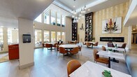 This sophisticated single-floor communal lounge offers a generously proportioned, open-plan interior designed with a refined modern aesthetic. The space is defined by its dramatic double-height ceilings, which flood the interior with natural light through expansive floor-to-ceiling windows and glass doors that frame views of an inviting outdoor pool and patio area. Large-format light gray tile flooring extends throughout, lending a clean, cohesive foundation to the space.

The main gathering area is anchored by multiple round and rectangular marble-topped tables paired with warm cognac leather chairs, accommodating both intimate and larger group settings. A striking cluster of glass globe pendant lights cascades from the ceiling, serving as a central design focal point. Along one wall, a sleek linear fireplace is framed by herringbone-patterned wall paneling and flanked by tall, dark-toned built-in shelving units displaying artful sculptures and decorative objects.

Adjacent areas include a tufted banquette seating section alongside a bold abstract art piece, and a billiards table nestled beneath an upper-level mezzanine with metal railings. A separate lounge nook near the entrance features plush velvet accent chairs. Large glass doors provide direct access to the outdoor amenity areas, seamlessly blending indoor and outdoor living.