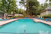 This outdoor pool area offers a welcoming retreat defined by its expansive, crystal-clear swimming pool with a classic rectangular layout and brick-edged coping. The pool is divided by a lane line, accommodating both recreational swimmers and lap swimmers alike. Surrounding the pool is a generous poured-concrete and brick sundeck furnished with numerous blue lounge chairs and dark wood-framed seating, providing ample space for relaxation and sunbathing. Bright yellow patio umbrellas are strategically positioned around the deck, offering shaded gathering spots for groups. The entire pool area is enclosed by a wrought iron safety fence, ensuring a secure and well-defined space. A charming single-story pool house with French doors and a hip roof sits directly adjacent to the pool, providing convenient access to amenities and facilities. The backdrop is dominated by towering mature pine and hardwood trees that create a lush, green canopy, lending the space a serene, nature-immersed atmosphere. Potted plants and manicured shrubbery along the fence line add additional color and landscaping detail. The overall ambiance is that of a well-maintained, resort-style outdoor amenity space ideal for community recreation and leisure.
