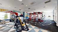 Located at 24770 E Applewood Cir in Aurora, CO, this fitness center offers a generously sized, single-floor exercise space designed to support a wide range of workout styles. The room is anchored by a bold, red multi-functional training rig equipped with cable attachments, TRX suspension straps, and a heavy punching bag, positioned at the center of a dark rubberized performance flooring zone featuring agility and circuit training markers. Surrounding this central area, the space transitions to a striking geometric, triangular-patterned carpet in blue, grey, and white tones, where a variety of Life Fitness selectorized strength machines and a dedicated free weights section with a full dumbbell rack are arranged for targeted resistance training. A separate cardio section along the windowed perimeter houses spin bikes, elliptical machines, and a treadmill, benefiting from natural light and outdoor views of the pool area, complemented by ceiling fans overhead. Recessed lighting throughout keeps the space bright and welcoming. The walls are adorned with vivid, large-scale Colorado landscape murals — depicting mountains, trees, and wildlife in a colorful, modern style — adding an energetic and regionally inspired ambiance. Wall-mounted flat-screen TVs provide additional entertainment throughout the facility.