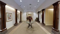 This single-floor space presents a sophisticated and formally designed lobby that immediately conveys a sense of refinement and attention to detail. The entryway is anchored by a central round glass-top table with a mirrored base, adorned with a striking floral arrangement in a deep burgundy vase, setting an elegant tone upon arrival. Flanking the central corridor are pairs of rich, dark wood columns with stone bases, adding a stately architectural character to the space. The walls are finished in a clean, neutral white, providing a bright backdrop that is complemented by soft recessed lighting overhead. Framed artwork decorates the walls on either side, adding visual interest and a curated aesthetic. A patterned area rug with a swirling motif grounds the central gathering area and adds warmth underfoot. The corridor leads gracefully through an arched passageway deeper into the building, hinting at additional spaces beyond. A classic upholstered accent chair is positioned thoughtfully near one of the columns, offering a welcoming resting spot. The overall ambiance is formal yet inviting, blending traditional design elements with tasteful décor to create an environment that feels both impressive and comfortable.