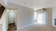 This two-floor space presents a straightforward, functional layout with a neutral color palette throughout. The main living area is finished with light-toned carpeting underfoot and features white walls that contribute to a bright, open feel. Vertical blinds cover a large window, allowing natural light to filter into the room while maintaining privacy. Exposed ductwork is visible near the ceiling in the living area, adding an industrial touch to the otherwise simple aesthetic.

A transition in flooring marks the shift from the carpeted living area to an adjoining hallway, where dark hardwood-style flooring provides a contrasting visual element. This corridor leads to additional areas of the space, with a doorway visible in the background hinting at further rooms beyond. The hallway is equipped with a ceiling light fixture, ensuring adequate illumination in transitional areas.

The walls are kept minimal with only essential fixtures such as outlets and switches mounted at standard heights. The overall design is clean and uncluttered, offering a neutral canvas adaptable to various furnishing styles. The two-floor configuration provides additional living or storage space, enhancing the overall functionality of this unit.