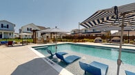 Located at 1392 E Bonds Ranch Rd in Fort Worth, Texas, this outdoor community pool area is a well-appointed recreational amenity designed for relaxation and social gathering. The centerpiece is a generously sized rectangular swimming pool with crystal-clear blue water and a mosaic tile border, complemented by a separate smaller wading or spa pool situated nearby. The expansive concrete sundeck surrounds the pool on all sides, providing ample space for lounging on the numerous blue chaise lounge chairs arranged throughout the area. Bold blue-and-white striped oversized umbrellas and standard patio umbrellas offer shade at various points across the deck. A dedicated dining and seating area shaded by a classic wooden pergola is positioned at one end of the pool, furnished with tables and chairs for casual outdoor meals and gatherings. On the opposite side, a cozy pod-style canopy daybed with blue cushions provides a private retreat. A neatly maintained turf lawn area adjacent to the pool features cornhole boards and additional seating, ideal for lawn games and socializing. The space is enclosed by a wrought iron fence with landscaped plantings, offering a sense of privacy while maintaining open views of the surrounding residential neighborhood under the open Texas sky.