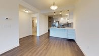 This single-floor, one-bedroom, one-bathroom unit offers a clean, modern aesthetic throughout. The open-concept living and dining area features dark hardwood-style flooring, crown molding, and a ceiling fan, creating a comfortable and well-finished space. Natural light enters through large windows, adding warmth to the neutral white walls and bright trim. The kitchen flows seamlessly from the living area and is outfitted with white shaker-style cabinetry, light quartz countertops, a gray subway tile backsplash, stainless steel appliances including a range and refrigerator, and a peninsula with pendant lighting overhead — ideal for casual dining or additional prep space. The bedroom is carpeted in soft gray and receives ample natural light from a large window with exterior views. The bathroom is thoughtfully designed with accessibility in mind, featuring a tub-shower combination with brushed nickel grab bars, white subway tile surround, large-format floor tile, and a floating-style vanity with a white cabinet base, stone countertop, and a framed mirror flanked by sconce lighting.