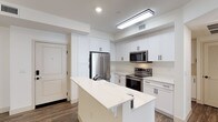This single-floor space showcases a thoughtfully designed kitchen that serves as its centerpiece. The kitchen is finished in a clean, all-white palette, featuring floor-to-ceiling cabinetry with matte black hardware that provides a subtle contrast to the bright surroundings. Ample upper and lower cabinet storage lines the walls, offering generous organization potential. The countertops are finished in white quartz with subtle veining, extending seamlessly across the workspace and onto a central kitchen island, which provides additional prep space and casual seating potential. Stainless steel appliances, including a French door refrigerator, an electric range, and an over-the-range microwave, lend a polished, contemporary look. Recessed and flush-mount ceiling lighting ensures the space is well-lit throughout. Wood-look flooring runs through the kitchen, adding warmth to the otherwise crisp aesthetic. The entry door is visible from the kitchen, suggesting an open, accessible layout. Overall, the space presents a clean, modern, and functional environment with a cohesive design scheme.