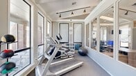 This fitness center in Lawton, Oklahoma offers a thoughtfully designed exercise room that maximizes its cozy footprint with an efficient and functional layout. The space is characterized by its bright, airy atmosphere, thanks to large windows lining the walls that flood the room with natural light and provide direct views of an outdoor pool area — creating an inviting backdrop for workouts.

The exercise room features a vaulted ceiling with exposed beam detailing and track lighting, lending a modern yet welcoming aesthetic. Neutral gray tones on the walls, accented by crisp white trim and framed wall panels, give the space a clean, polished look. Carpet flooring runs throughout, providing a comfortable surface underfoot.

Equipment includes treadmills, an elliptical cross-trainer, a stability ball, and a dedicated free weights area outfitted with dumbbells, resistance bands, and interlocking floor mats for stretching or floor exercises. A multi-tiered storage unit keeps smaller accessories neatly organized. A wall-mounted mirror in the free weights area enhances the sense of space and supports form-checking during exercise.

Glass panel partitions on one side of the room allow visual connectivity to an adjacent space, contributing to an open, inclusive feel. The overall ambiance is bright, clean, and motivating — well-suited for a focused fitness routine.