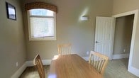 This single-floor space exudes a cozy and welcoming ambiance through its thoughtful design and warm neutral palette. The dining area serves as a notable feature, centered around a natural wood dining table accompanied by matching light wood chairs, creating a cohesive and casual yet refined atmosphere. A standout architectural detail is the arched window adorned with a mosaic tile accent above the blinds, allowing warm natural light to filter into the room and adding a touch of elegant character to the space. The walls are finished in a soft, earthy taupe tone that complements the warm wood tones of the furniture, contributing to the overall sense of comfort and calm. White painted trim and a classic paneled door add crisp contrast and a timeless quality to the interior. A wall-mounted light fixture provides additional illumination, while the neutral carpeted flooring ties the space together. The layout feels open and functional, offering a pleasant setting for everyday dining or casual gatherings. Overall, the space balances practical design with subtle architectural charm, making it both livable and visually appealing.
