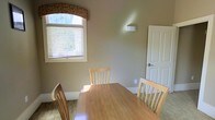 This single-floor space exudes a cozy and welcoming ambiance through its thoughtful design and warm neutral palette. The dining area serves as a notable feature, centered around a natural wood dining table accompanied by matching light wood chairs, creating a cohesive and casual yet refined atmosphere. A standout architectural detail is the arched window adorned with a mosaic tile accent above the blinds, allowing warm natural light to filter into the room and adding a touch of elegant character to the space. The walls are finished in a soft, earthy taupe tone that complements the warm wood tones of the furniture, contributing to the overall sense of comfort and calm. White painted trim and a classic paneled door add crisp contrast and a timeless quality to the interior. A wall-mounted light fixture provides additional illumination, while the neutral carpeted flooring ties the space together. The layout feels open and functional, offering a pleasant setting for everyday dining or casual gatherings. Overall, the space balances practical design with subtle architectural charm, making it both livable and visually appealing.