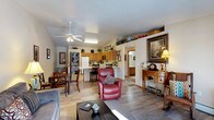 This single-floor home offers a thoughtfully designed open-concept layout that connects the living, dining, and kitchen areas into one cohesive, functional space. The living area features warm hardwood-style flooring that flows throughout, anchored by a comfortable seating arrangement and a ceiling fan that enhances the light, airy feel of the room. Vaulted ceilings in the living space add a sense of openness and volume, while natural light filters in to brighten the interior throughout the day. A centrally positioned kitchen island with a breakfast bar serves as a natural dividing point between the kitchen and the main living area, offering additional seating and prep space. The kitchen is outfitted with warm wood cabinetry, a full suite of appliances including a refrigerator and stove, and ample counter space for everyday use. The adjacent dining area comfortably accommodates a full dining table and chairs, making it ideal for meals and gatherings. Decorative shelving above the kitchen cabinets provides additional display space, adding a personalized touch to the home. A hallway visible from the main living area leads to additional rooms, suggesting a practical and well-organized floor plan. The overall ambiance is warm, welcoming, and well-suited for comfortable everyday living.