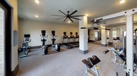 This single-level fitness center is thoughtfully designed to accommodate a comprehensive workout experience within a spacious, open layout. The space is organized into several distinct zones connected by a wide, carpeted corridor with structural columns and dark-trimmed doorways that lend a clean, modern aesthetic throughout.

The primary cardio area features a row of treadmills and elliptical machines, each equipped with individual monitor screens for entertainment. Oversized ceiling fans with broad blade spans provide airflow and contribute to the room's contemporary industrial character. Recessed lighting throughout ensures consistent illumination across all workout zones.

A dedicated free weights area includes a multi-tiered dumbbell rack and adjustable bench, with wall-mounted flat-screen televisions positioned for viewing during exercise. An additional strength training section houses cable and resistance machines, visible through the open floor plan.

Large windows and glass-panel entry doors in the free weights room allow natural light to filter in while offering views of the surrounding exterior. The neutral carpeted flooring and light-toned walls create a clean, professional environment.

Adjacent to the main fitness floor is a lounge and kitchenette space, featuring dark cabinetry, a stainless steel sink, beverage refrigerator, and decorative wall sconces. Natural light enters through double windows overlooking a landscaped outdoor area, and rustic wood stump side tables add a warm, informal touch to the seating area.