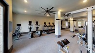 This single-level fitness center is thoughtfully designed to accommodate a comprehensive workout experience within a spacious, open layout. The space is organized into several distinct zones connected by a wide, carpeted corridor with structural columns and dark-trimmed doorways that lend a clean, modern aesthetic throughout.

The primary cardio area features a row of treadmills and elliptical machines, each equipped with individual monitor screens for entertainment. Oversized ceiling fans with broad blade spans provide airflow and contribute to the room's contemporary industrial character. Recessed lighting throughout ensures consistent illumination across all workout zones.

A dedicated free weights area includes a multi-tiered dumbbell rack and adjustable bench, with wall-mounted flat-screen televisions positioned for viewing during exercise. An additional strength training section houses cable and resistance machines, visible through the open floor plan.

Large windows and glass-panel entry doors in the free weights room allow natural light to filter in while offering views of the surrounding exterior. The neutral carpeted flooring and light-toned walls create a clean, professional environment.

Adjacent to the main fitness floor is a lounge and kitchenette space, featuring dark cabinetry, a stainless steel sink, beverage refrigerator, and decorative wall sconces. Natural light enters through double windows overlooking a landscaped outdoor area, and rustic wood stump side tables add a warm, informal touch to the seating area.