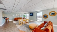 This pool lounge in Memphis, Tennessee, is a well-appointed communal space designed for relaxation and socializing. The open-plan layout is anchored by light wood-look flooring and soft sage-green walls, creating a fresh and inviting atmosphere throughout. A drop tile ceiling with recessed lighting keeps the space feeling bright and polished.

The lounge area is generously furnished with rich cognac leather sofas, accent chairs, and layered area rugs, offering multiple comfortable seating arrangements. A wall-mounted flat-screen TV serves as a central focal point, ideal for group gatherings. A large dining table with wooden chairs provides a dedicated space for casual meals or social activities. Decorative touches, including abstract wall art and round mirror accents, add personality to the space.

Large windows and glass doors flood the interior with natural light and frame views of the outdoor pool area and the Memphis skyline beyond, creating a seamless connection between indoor and outdoor living.

The kitchenette is a standout feature, boasting a striking gold penny-tile mosaic backsplash, dark countertops, a bar counter with stylish wooden barstools, white subway tile accents, gray cabinetry, and a full-size stainless steel refrigerator — all thoughtfully designed to support entertaining and everyday use.