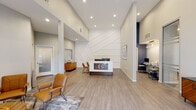 Located at 8965 E Florida Ave in Denver, Colorado, this single-floor professional office space combines modern design with a warm, welcoming atmosphere. The reception area serves as the focal point of the space, featuring a sleek, branded front desk with walnut wood accents and a striking geometric accent wall finished in crisp white. Soaring ceilings amplify the sense of openness, while recessed lighting throughout creates a clean, well-lit environment. Light-toned hardwood-style flooring flows seamlessly across the entire space, lending a cohesive and polished aesthetic. A dedicated waiting area offers comfortable cognac leather chairs arranged around a circular wood coffee table, complemented by natural light from nearby windows with roller shades. A self-service workstation station is conveniently integrated into the main area, accented by a bold navy blue wall. The space also includes a glass-paneled interior door that adds a layer of transparency and visual connectivity between areas. Colorado-inspired artwork adorns the walls, reinforcing a sense of local character. A warm wood credenza and decorative accessories complete the refined, professional look. The front entry features glass doors that invite natural light and offer views of the surrounding landscaped exterior, creating an inviting first impression for all visitors.