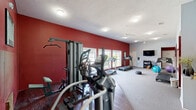 Located in Biloxi, Mississippi, this fitness center offers a well-organized and energetic workout environment designed to support a full range of exercise routines. The space is defined by its bold red accent walls, which contrast with light gray tones to create a motivating atmosphere throughout.

The exercise room is divided into two functional zones. The cardio area features multiple treadmills and an elliptical trainer, all positioned along a row of large windows that flood the space with natural light and offer views of the surrounding outdoor area. Ceiling fans provide additional airflow and comfort during workouts. A wall-mounted television and a punching bag stand add variety to the cardio section.

The strength training zone transitions to rich, dark hardwood flooring and is equipped with a multi-station cable machine, a weight bench, and additional resistance equipment. A large mirror is mounted on the wall, enhancing the sense of space and allowing users to monitor their form. Stability balls and free weights round out the available equipment.

Textured carpet covers the main cardio floor area, while the strength zone's hardwood flooring provides a distinct and practical surface. Ceiling-mounted light fixtures ensure even illumination throughout. The space is accessible via two entry doors and is well-ventilated, making it a functional and inviting facility for daily fitness use.