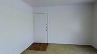 This single-floor space presents a straightforward and functional layout characterized by clean, white walls that create a bright, neutral atmosphere throughout. The entry area features a section of dark hardwood flooring that transitions into wall-to-wall carpet, clearly delineating the entryway from the main living area. The solid white entry door is equipped with dual locking mechanisms, providing a secure point of access.

The walls are smooth and painted in a crisp white, contributing to an open and airy feel despite the modest scale of the space. Electrical outlets are positioned along the walls, offering practical placement for furnishings and appliances. The flooring combination of hardwood at the entry and carpet in the main area adds a degree of visual interest while maintaining a practical design.

The overall ambiance of the space is clean and uncluttered, presenting a blank canvas that offers flexibility for a variety of furnishing arrangements. The minimalist design makes it easy to envision how the space could be personalized and utilized effectively.