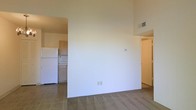 This single-floor space showcases a practical, open-concept layout that seamlessly connects the living area to the kitchen. The living space is finished with wall-to-wall carpeting, while the kitchen transitions to tile flooring, creating a clear and functional distinction between the two areas. The kitchen is equipped with standard cabinetry in a warm wood tone, a white refrigerator, and a countertop workspace, offering efficient use of the available area.

The walls throughout are painted in a clean, bright white, contributing to an airy and neutral ambiance that provides a versatile backdrop for a variety of furnishing styles. A central dividing wall separates the main living space from the kitchen without fully enclosing either area, maintaining an open and connected feel. A ceiling vent ensures climate control, and standard electrical outlets are conveniently positioned along the baseboards.

A doorway visible to the right of the space suggests access to additional rooms or a hallway, indicating further functionality beyond the main living and kitchen areas. Overall, the space presents a clean, straightforward layout suited to everyday living.