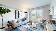 This single-floor unit presents a thoughtfully designed open-concept layout that seamlessly connects the kitchen, dining, and living areas. The kitchen is outfitted with light wood upper cabinetry, white lower cabinets, a white subway tile backsplash, stainless steel appliances, and a marble-topped island with bar seating — ideal for casual dining. Light wood-toned flooring runs throughout the main living areas, contributing to a warm, cohesive aesthetic.

The living room is bright and inviting, anchored by a large window fitted with horizontal blinds that frame outdoor views and allow generous natural light to fill the space. The room accommodates a comfortable seating arrangement alongside a media console area with ample wall space for artwork.

The bedroom features plush carpet flooring, a dedicated wall shelf, and direct access to a spacious walk-in closet with built-in rod storage. The room is well-proportioned, offering flexibility in furniture arrangement.

The bathroom is finished in a clean, modern style with a wide white vanity, quartz countertop, undermount sink, and a backlit mirror that enhances the bright, airy feel of the space. Overall, the unit maintains a consistent contemporary design with a neutral, welcoming palette throughout.