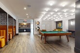 This thoughtfully designed amenity floor blends modern aesthetics with warm, inviting materials throughout its distinct zones. Wood-look tile flooring flows consistently across the entire level, unifying the space while complementing contrasting design elements. A dramatic double-height atrium anchors the layout, framed by exposed brick walls, floor-to-ceiling industrial-style windows, and warm wood ceiling accents that flood the area with natural light. A mezzanine level with a metal mesh railing overlooks this central gathering space, which features curated lounge seating, bold accent furniture, and oversized planters.

The game room showcases a full-size billiards table beneath a striking cluster of globe pendant lights, with decorative lattice screens and colorful wall art adding character. Adjacent to this area, a business nook provides built-in shelving, cabinetry, and workspace essentials. A café-style bar counter with a wood surface, green tile backsplash, and brass pendant lighting serves as a social hub. Private dining booths with mustard-yellow upholstery, patterned wallcoverings, and starburst pendants offer an intimate setting. A utility and service alcove with dark cabinetry and open shelving completes the floor.