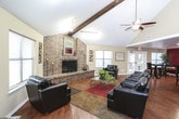 This single-floor space functions as a well-appointed common area or clubhouse lounge, thoughtfully divided into distinct zones that serve different gathering needs. The main lounge area commands attention with its dramatic vaulted ceilings accented by exposed wood beams and dual ceiling fans, creating an open and airy atmosphere. Track lighting runs throughout, supplementing the abundant natural light that streams in through multiple large windows fitted with blinds.

A standout feature is the full brick fireplace with a raised hearth, serving as a striking architectural focal point in the lounge. Dark leather sofas and armchairs are arranged around a glass-top coffee table atop layered area rugs, offering a comfortable and inviting seating arrangement. Decorative wall art and greenery add warmth throughout.

Transitioning toward the dining area, bold crimson accent walls create a visual contrast against the neutral tones of the rest of the space. High-top pub tables paired with dark wood bar stools provide casual seating, while a wall-mounted flat-screen television adds a functional entertainment element. Rich hardwood flooring runs seamlessly throughout, tying the entire space together with a cohesive, polished aesthetic. A kitchenette area is visible in the background, enhancing the space's utility.
