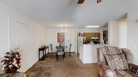 This single-floor residence offers an open-concept layout that effortlessly connects the main living spaces, creating a cohesive and functional environment. The living area features carpeted flooring and is furnished with a classic floral sofa, complemented by a ceiling fan overhead. Adjacent to the living area, the dining space is defined by a decorative chandelier that adds a touch of elegance, centered above a glass-top dining table with sleek black metal chairs. A piece of wall art adds a personal, artistic accent to the room. The kitchen is accessible via a peninsula-style counter that serves as a natural divider between the dining and cooking areas. Warm wood-toned cabinetry lines the kitchen walls, providing ample storage, while a white refrigerator and standard appliances complete the space. A fluorescent light fixture ensures bright, practical illumination in the kitchen. The transition between the tiled entryway and the carpeted main areas subtly delineates the different zones within the home. The textured ceiling throughout adds a classic residential character, and the overall layout promotes an easy flow between spaces, making this residence both practical and inviting.