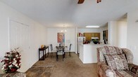 This single-floor residence offers an open-concept layout that effortlessly connects the main living spaces, creating a cohesive and functional environment. The living area features carpeted flooring and is furnished with a classic floral sofa, complemented by a ceiling fan overhead. Adjacent to the living area, the dining space is defined by a decorative chandelier that adds a touch of elegance, centered above a glass-top dining table with sleek black metal chairs. A piece of wall art adds a personal, artistic accent to the room. The kitchen is accessible via a peninsula-style counter that serves as a natural divider between the dining and cooking areas. Warm wood-toned cabinetry lines the kitchen walls, providing ample storage, while a white refrigerator and standard appliances complete the space. A fluorescent light fixture ensures bright, practical illumination in the kitchen. The transition between the tiled entryway and the carpeted main areas subtly delineates the different zones within the home. The textured ceiling throughout adds a classic residential character, and the overall layout promotes an easy flow between spaces, making this residence both practical and inviting.