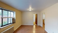 This single-floor space offers a straightforward and functional layout with a clean, understated aesthetic. The main living area features warm hardwood flooring that extends throughout, complemented by light yellow-toned walls and white trim that create a bright, welcoming atmosphere. A flush-mount ceiling light provides overhead illumination, while a window fitted with a wall-mounted air conditioning unit allows for natural light and ventilation. A traditional radiator along the base of one wall indicates a classic heating system. The room connects seamlessly to additional spaces through two open passageways — one leading to what appears to be a bedroom or secondary room through a classic wooden door, and another opening directly into a kitchen area. The kitchen, visible from the main room, features cabinetry and a countertop, suggesting a compact yet practical cooking space. The overall layout is open and easy to navigate, with neutral finishes throughout that provide a versatile backdrop. The space maintains a simple, no-frills character suited to practical, everyday living.
