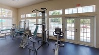 This single-floor fitness center is thoughtfully designed to support a full range of workout routines. The space is generously sized, with durable dark rubber flooring throughout that provides both safety and functionality for exercise. Neutral, light-toned walls and high ceilings contribute to an open, airy atmosphere, while large windows and glass-paneled doors along multiple walls flood the interior with abundant natural light and offer views of the surrounding outdoor areas, including a pool and landscaped grounds.

The cardio zone features multiple treadmills and elliptical machines, all positioned in front of a large wall-to-wall mirror that enhances the sense of depth and allows users to monitor their form. Flat-screen televisions are mounted for entertainment during workouts. The strength-training area is well-appointed with a variety of cable and weight machines, a pull-up and dip station, a stationary recumbent bike, and additional resistance equipment, providing options for targeting multiple muscle groups.

Overhead structural beams add an architectural element to the space, while ample room between equipment ensures comfortable movement throughout. Direct access to the outdoor area via French-style doors further enhances the facility's connection to the surrounding environment.