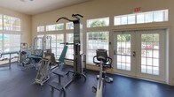 This single-floor fitness center is thoughtfully designed to support a full range of workout routines. The space is generously sized, with durable dark rubber flooring throughout that provides both safety and functionality for exercise. Neutral, light-toned walls and high ceilings contribute to an open, airy atmosphere, while large windows and glass-paneled doors along multiple walls flood the interior with abundant natural light and offer views of the surrounding outdoor areas, including a pool and landscaped grounds.

The cardio zone features multiple treadmills and elliptical machines, all positioned in front of a large wall-to-wall mirror that enhances the sense of depth and allows users to monitor their form. Flat-screen televisions are mounted for entertainment during workouts. The strength-training area is well-appointed with a variety of cable and weight machines, a pull-up and dip station, a stationary recumbent bike, and additional resistance equipment, providing options for targeting multiple muscle groups.

Overhead structural beams add an architectural element to the space, while ample room between equipment ensures comfortable movement throughout. Direct access to the outdoor area via French-style doors further enhances the facility's connection to the surrounding environment.