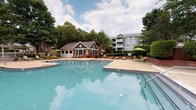 Located at 8810 Autumn Woods Dr in Raleigh, NC, this single-level outdoor amenity space is designed for relaxation and social gathering. The centerpiece is a generously sized resort-style swimming pool with clear blue water, surrounded by a spacious concrete deck lined with lounge chairs and shade umbrellas — ideal for sunbathing or unwinding outdoors. A dedicated pool house with large windows and a charming roofline provides convenient support facilities poolside. The covered patio area features classic brick construction accented by crisp white columns and herringbone-patterned brick flooring, offering a shaded retreat with outdoor seating arrangements perfect for socializing. Adjacent to the patio, a built-in outdoor kitchen equipped with stainless steel grills sits atop a stone counter, making al fresco dining effortless. Mature trees and manicured shrubbery, including crape myrtles and sculpted hedgerows framed by low brick retaining walls, provide natural privacy and a serene, park-like atmosphere throughout the grounds. The overall ambiance is welcoming and well-maintained, blending traditional brick architectural details with lush greenery to create an inviting community outdoor living experience.