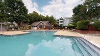 Located at 8810 Autumn Woods Dr in Raleigh, NC, this single-level outdoor amenity space is designed for relaxation and social gathering. The centerpiece is a generously sized resort-style swimming pool with clear blue water, surrounded by a spacious concrete deck lined with lounge chairs and shade umbrellas — ideal for sunbathing or unwinding outdoors. A dedicated pool house with large windows and a charming roofline provides convenient support facilities poolside. The covered patio area features classic brick construction accented by crisp white columns and herringbone-patterned brick flooring, offering a shaded retreat with outdoor seating arrangements perfect for socializing. Adjacent to the patio, a built-in outdoor kitchen equipped with stainless steel grills sits atop a stone counter, making al fresco dining effortless. Mature trees and manicured shrubbery, including crape myrtles and sculpted hedgerows framed by low brick retaining walls, provide natural privacy and a serene, park-like atmosphere throughout the grounds. The overall ambiance is welcoming and well-maintained, blending traditional brick architectural details with lush greenery to create an inviting community outdoor living experience.