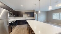 The Sierra floor plan at 800 Railyard Loop in Grand Junction, Colorado presents a thoughtfully designed 2-bedroom, 2-bathroom single-floor residence with a cohesive modern aesthetic throughout. The open-concept kitchen serves as a centerpiece, featuring dark espresso cabinetry, a large central island with a bright white quartz countertop, stainless steel appliances including a refrigerator, over-the-range microwave, and electric range, all complemented by light wood-style flooring and pendant lighting. The adjacent living area offers neutral-toned walls, plush carpeting, and access to an exterior door, allowing natural light to flow in through a large window. Both bedrooms are finished with soft carpet, neutral wall tones, and windows that bring in natural light. The primary bedroom includes two closets—one being a spacious walk-in with wire shelving—and direct access to its private bathroom. The bathrooms feature dark vanity cabinetry, white quartz countertops, and a tub/shower combination with chrome fixtures and light wood-style flooring. A hallway connects the space efficiently, and the overall design maintains a clean, contemporary feel throughout.